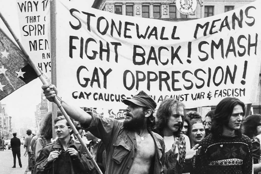 A black and white photo of a protest with participants holding banners. One prominent banner reads, "STONEWALL MEANS FIGHT BACK! SMASH GAY OPPRESSION!" 