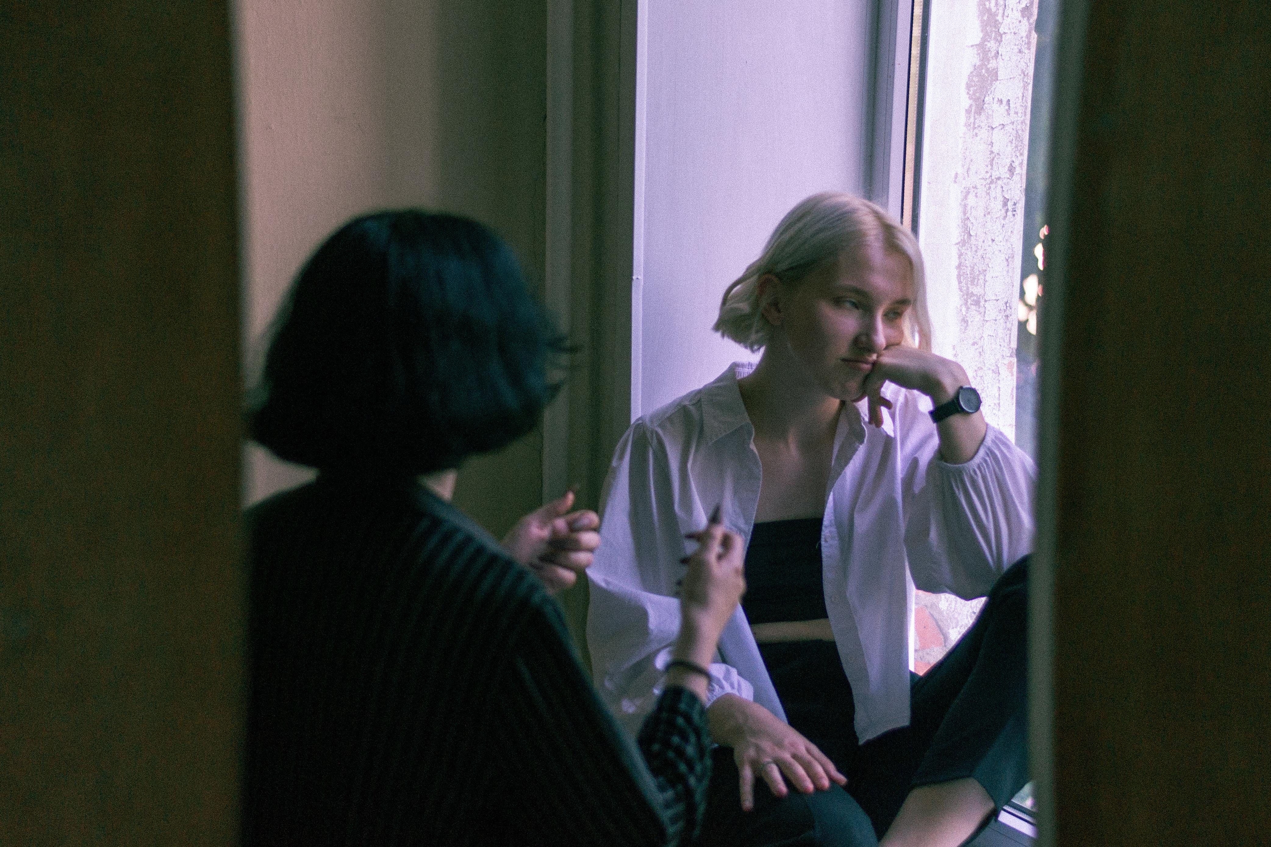 A photo capturing a conversation between two white women, taken through a doorway. The person in the foreground is seen from the back, with short dark hair, wearing a striped garment. Facing them is a person with short blonde hair, dressed in a white shirt, resting their chin on their hand with a pensive expression