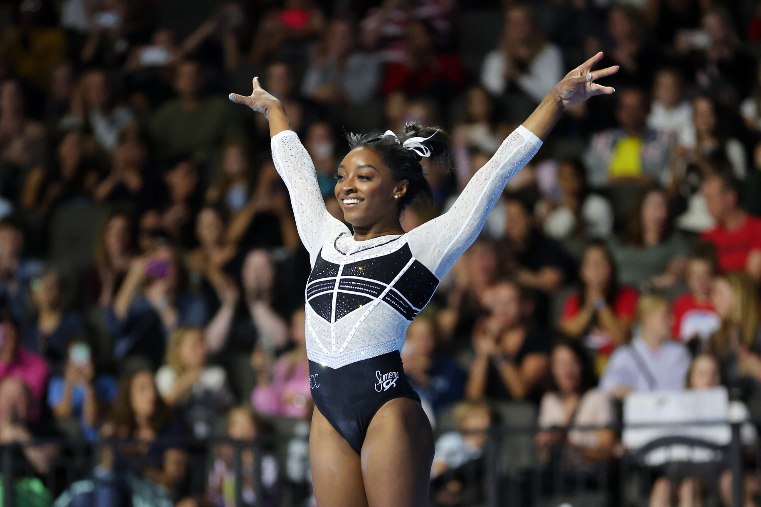 A photo of a female gymnast Simone Biles in mid-performance, extending her arms wide with a joyful expression. She's wearing a leotard with white and dark designs resembling a starry night. The background is filled with a blurred audience in a gymnastics arena.
